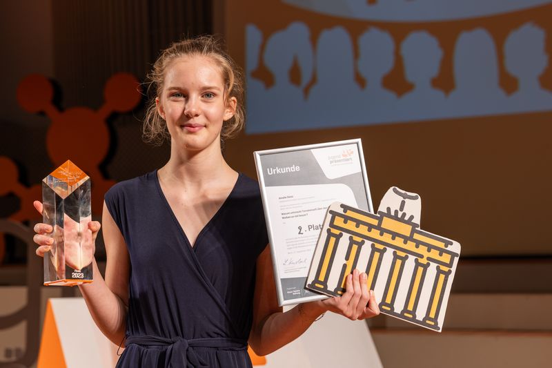 Die Schülerin Amelie Deisz hält ihren Pokal und ihre Urkunde für den zweiten Platz in die Kamera. Zudem hat sie ein Pappbild mit einer Brandenburger-Tor-Illustration in der Hand.